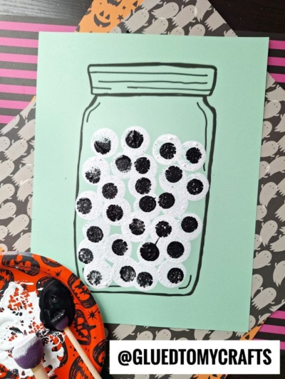 A drawing of a stamped eyeball jar on green paper is filled with black and white painted circles resembling eyeballs. Halloween-themed papers and paint supplies are nearby. @GLUEDTOMYCRAFTS is labeled at the bottom.