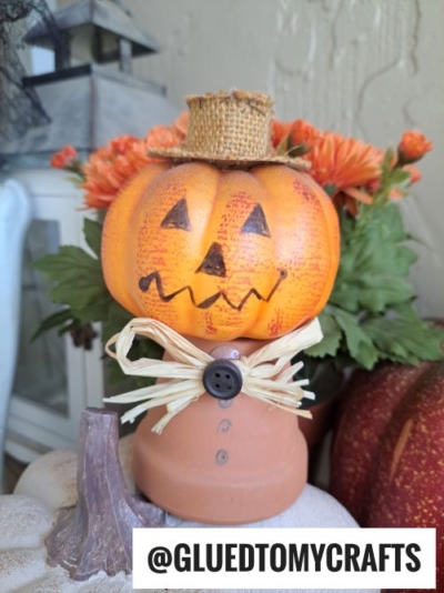 A pumpkin-headed scarecrow decoration with a burlap hat and raffia bow stands on a terra cotta pot, surrounded by orange flowers and pumpkins. The text @GLUEDTOMYCRAFTS appears at the bottom—perfect for terra cotta pot crafts lovers.