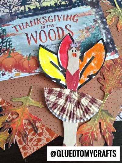 A handmade craft stick turkey ballerina with a cupcake liner skirt, colorful paper feathers, and a drawn face sits on autumn-themed paper and a Thanksgiving in the Woods book. Text: @GLUETOMYCRAFTS.