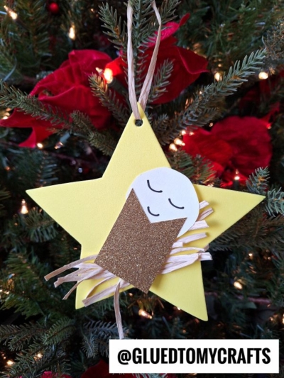 A yellow baby Jesus Christmas star ornament made of paper and glitter, featuring a sweet baby Jesus at its center, hangs on a Christmas tree adorned with red flowers and white lights. @GLUEDTOMYCRAFTS is written at the bottom.