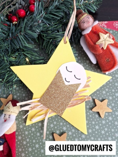 A handmade Christmas ornament shaped like a yellow baby Jesus Christmas star features a paper and straw baby Jesus, surrounded by festive decorations like felt angel and Santa figurines, plus gold stars. @gluedtomycrafts is written at the bottom.