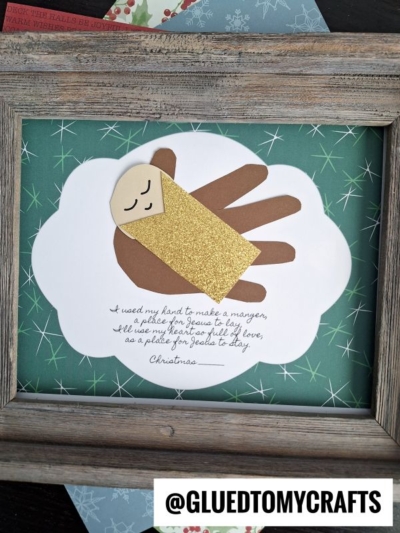 Framed craft featuring a Handprint Baby Jesus made with a brown handprint as the manger and gold glitter for the swaddle, lying on a white cloud shape with handwritten text below, all set against a green patterned background.