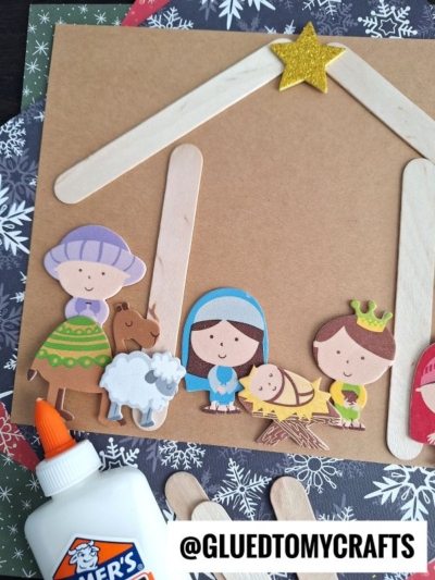 A nativity scene kid craft featuring paper cutouts of Mary, Joseph, baby Jesus, wise men, animals, and a popsicle stick stable with a yellow star. A bottle of glue sits nearby to help kids assemble their festive creation.