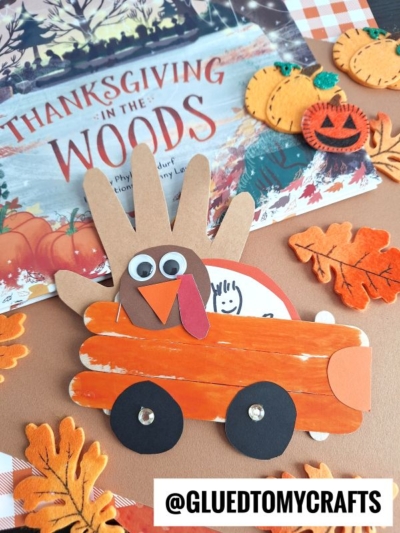 A popsicle stick Thanksgiving truck, decorated with googly eyes and paper feathers, sits on orange wheels. Autumn leaves, felt pumpkins, and a Thanksgiving in the Woods book surround it. @GLUEDTOMYCRAFTS is shown below.