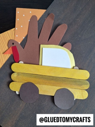 A turkey made from a handprint and paper pieces is sitting in a popsicle stick Thanksgiving truck with brown wheels. The image features craft paper in the background and the handle @GLUEDTOMYCRAFTS at the bottom right.