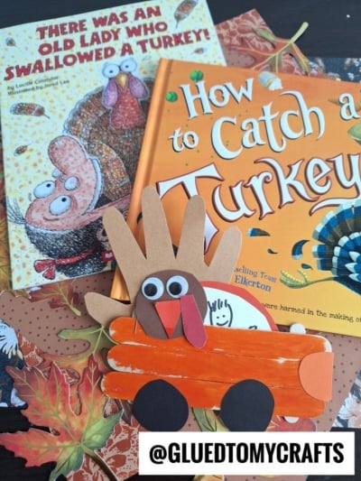 Two children’s books, There Was an Old Lady Who Swallowed a Turkey! and How to Catch a Turkey, lie on a table with autumn leaves. A popsicle stick Thanksgiving truck holds a handprint turkey craft in front. @GLUEDTOMYCRAFTS text is at the bottom.