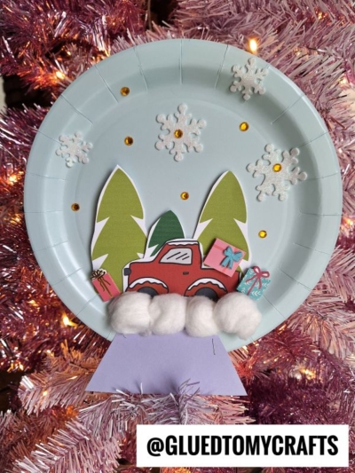 Paper plate craft featuring a festive red truck Christmas snow globe scene, with gift boxes, trees, cotton balls for snow, snowflake stickers, and gold dots—all beautifully displayed on a pink tinsel tree. Text at the bottom reads @GLUEDTOMYCRAFTS.