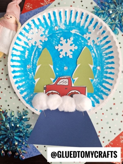 A red truck Christmas snow globe craft made from a painted paper plate, decorated with paper trees, a red car, white cotton snow, and snowflake cutouts, set on festive holiday paper. @GLUEDTOMYCRAFTS is at the bottom.