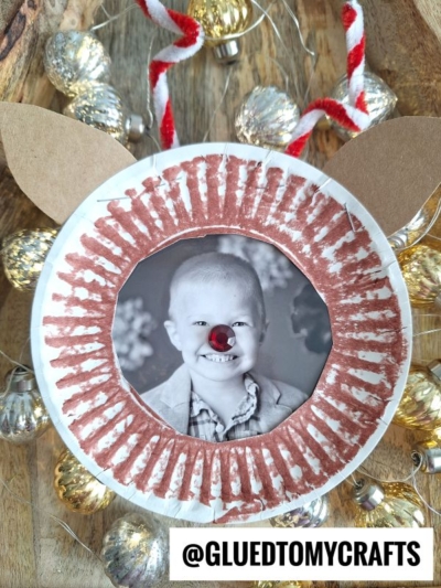 A festive paper plate reindeer picture frame features a child’s black-and-white photo at the center with a red pompom nose, brown painted lines, cardboard ears, and holiday lights and candy cane garland. Text reads @GLUEDTOMYCRAFTS.