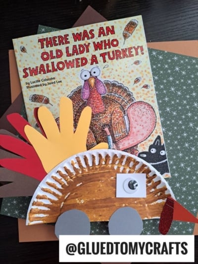 A children’s book titled “There Was an Old Lady Who Swallowed a Turkey!” is next to a handmade paper plate turkey craft, made from colored paper and a googly eye, with colorful paper sheets and toy cars in the background.