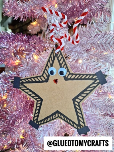 A reindeer star-shaped ornament with googly eyes, a red gem nose, and patterned borders hangs on a pink Christmas tree. It’s attached with a red and white twisted string. Text reads @GLUEDTOMYCRAFTS.