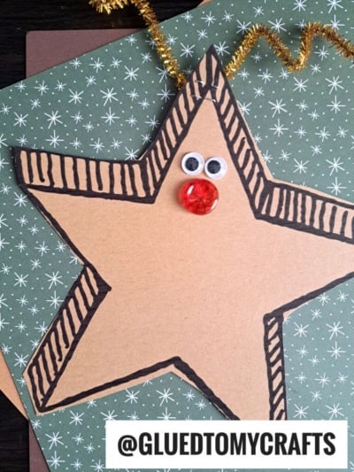 A reindeer star craft made from brown paper, outlined in black, features googly eyes, a red gem nose, and gold pipe cleaner antlers. It’s displayed on green star-patterned paper with @GLUEDTOMYCRAFTS at the bottom.