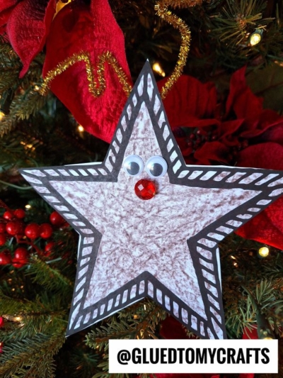 A handmade reindeer star ornament with googly eyes and a red gem nose hangs on a Christmas tree. The star is outlined in black with white accents and topped with gold pipe cleaner for festive charm.