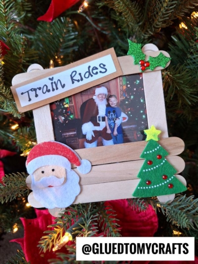 A holiday photo frame decorated with felt Santa, tree, and holly holds a picture of a child with Santa Claus. The frame says Train Rides and hangs on a popsicle stick Christmas tree stand. @GLUETOMYCRAFTS is at the bottom.