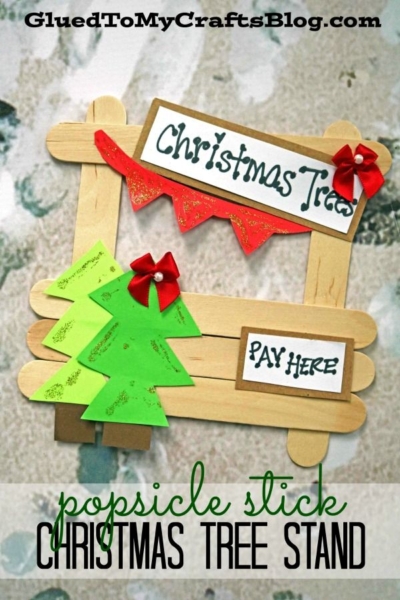 A popsicle stick frame decorated with paper Christmas trees, bows, and signs reading Christmas Trees and Pay Here, perfectly represents a festive popsicle stick Christmas tree stand craft. Text overlay says popsicle stick CHRISTMAS TREE STAND.