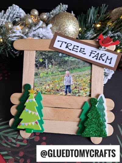 A handmade wooden frame decorated with foam Christmas trees displays a photo of a child outdoors. Above the frame is a sign reading “TREE FARM,” and beside it sits a popsicle stick Christmas tree stand amid snowy, festive holiday decor.