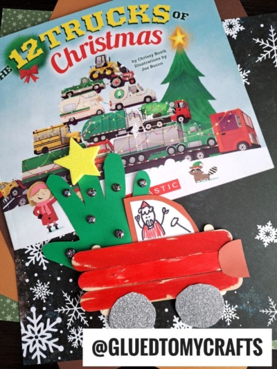 A children’s book titled The 12 Trucks of Christmas is shown with a popsicle stick christmas truck craft made from craft sticks, foam, and paper shaped like a red truck, next to the book. The background is decorated with festive paper.