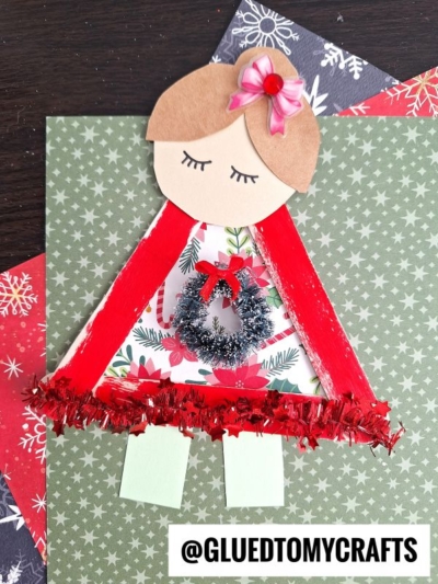 A handmade paper angel craft with brown hair, a red ribbon cape, tinsel, a festive wreath, and closed eyes, displayed on a green patterned background—perfect to pair with an ugly sweater craft. The handle @GLUEDTOMYCRAFTS is at the bottom.