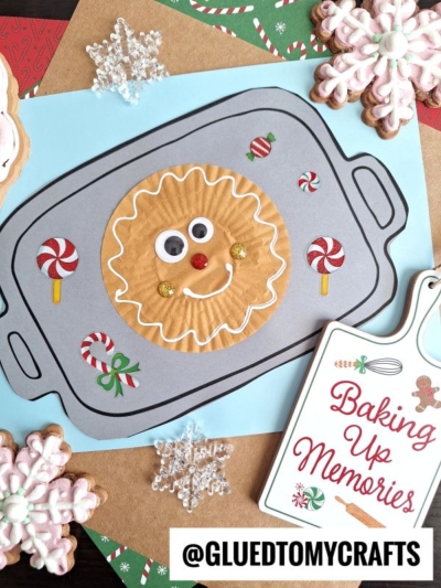 A delightful cookie sheet craft featuring a smiling gingerbread face made from a cupcake liner on paper shaped like a Christmas cookie sheet, adorned with candy stickers, snowflake decorations, and a cheerful Baking Up Memories sign nearby.