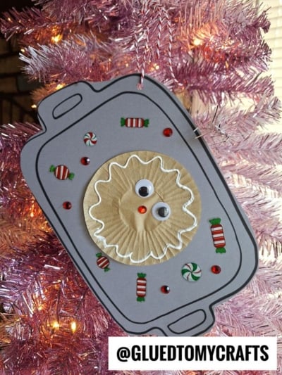 A handmade Christmas ornament shaped like a paper Christmas cookie sheet hangs on a pink tree. It features a ruffled paper cup decorated as a gingerbread cookie face, with candy and gem stickers around it. @GLUEDTOMYCRAFTS text is at the bottom.