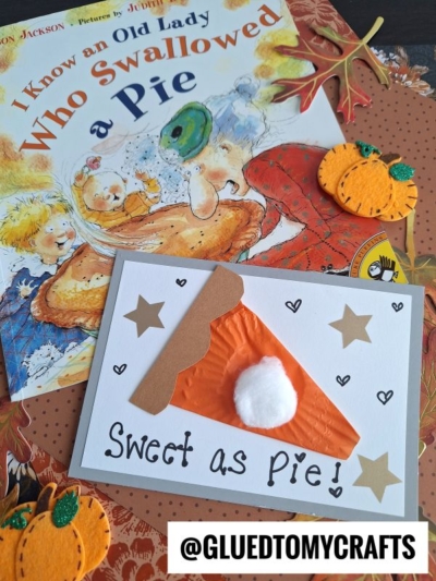 A handmade card with a slice of pie crafted from orange paper and a cotton ball sits on autumn-themed stationery, beside the book I Know an Old Lady Who Swallowed a Pie and cupcake liner pumpkin pie crafts.