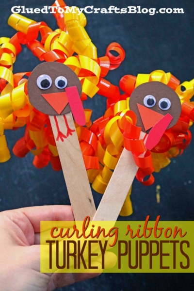 Two handmade curling ribbon turkey puppets made from craft sticks, curly yellow and orange ribbon, brown paper heads, googly eyes, and paper beaks are held up against a dark background. Text reads: curling ribbon TURKEY PUPPETS.