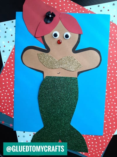 A paper mermaid craft on a blue background, made with colored paper shapes, googly eyes, a red nose gem, a black bow on the hair, a gold glitter top, and a green glitter tail—perfect to disguise the gingerbread man! The handle @GLUEDTOMYCRAFTS is shown.