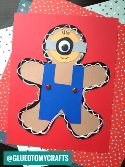 A gingerbread man craft made from brown paper is cleverly disguised as a Minion, featuring a single eye, blue overalls, large buttons, and white icing details along the edges, all set against a red background.