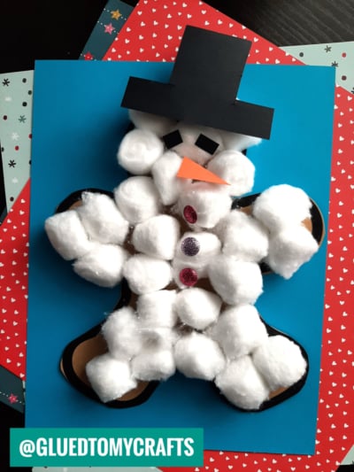 A gingerbread man-shaped craft is decorated with cotton balls to disguise the gingerbread man as a snowman. It features a black paper top hat, orange paper nose, black eyes, and glittery buttons on colored paper.