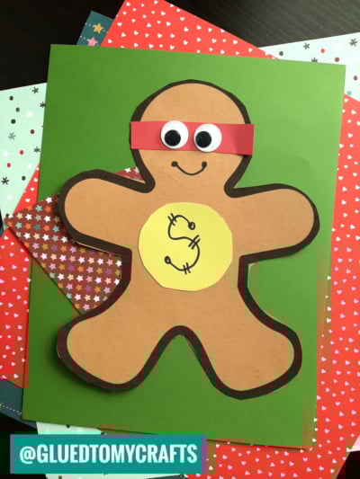 A paper craft gingerbread superhero in disguise, with a red mask, googly eyes, and a yellow circle with a dollar sign on its chest, placed on green and star-patterned papers.