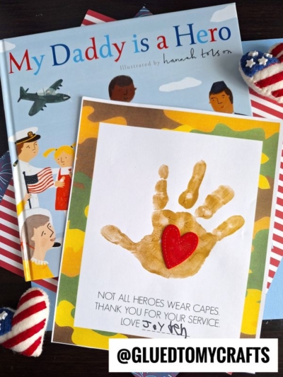 A handmade card with a child’s tan handprint and red heart, reading “Not all heroes wear capes. Thank you for your service. Love Joey,” is displayed beside My Daddy is a Hero and patriotic heart decorations.