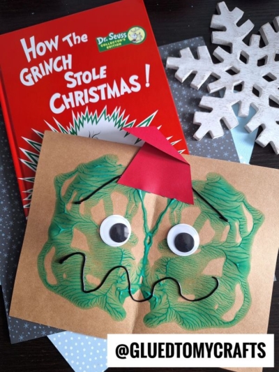 A children’s book titled “How the Grinch Stole Christmas!” is shown with a Paint Splat Grinch face craft made from paint, googly eyes, and a red paper hat. A wooden snowflake and a @GLUEDTOMYCRAFTS sign are nearby.