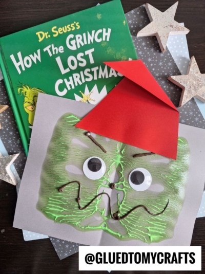 A Dr. Seuss How The Grinch Lost Christmas! book next to a Paint Splat Grinch face craft made with green paint, googly eyes, a red paper hat, and brown string, displayed on a table with paper stars. @GLUEDTOMYCRAFTS watermark at the bottom.