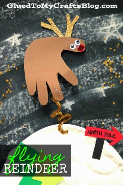 A paper flying reindeer, crafted from a brown handprint with googly eyes, a red pom-pom nose, and gold antlers, soars above a snow-covered North Pole sign on a starry black background.