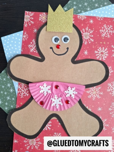 A paper gingerbread princess with a gold crown, googly eyes, red nose, and a pink cupcake liner dress decorated with white snowflakes and red gems is placed on festive snowflake-patterned papers. Perfect for making paper gingerbread princesses!.