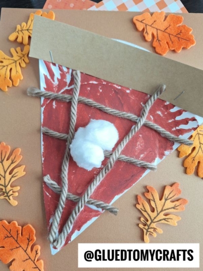 A kids craft of a paper plate apple pie slice made from painted paper, yarn crust, and cotton whipped cream, surrounded by felt orange leaves. The Instagram handle @gluedtomycrafts is at the bottom.