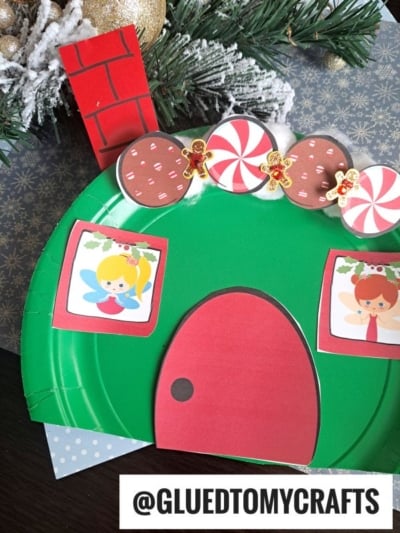 A green paper plate Christmas fairy house with a red door, illustrated windows, chimney, candy, and gingerbread designs sits beside a snowy pine branch. @GLUEDTOMYCRAFTS is written at the bottom.