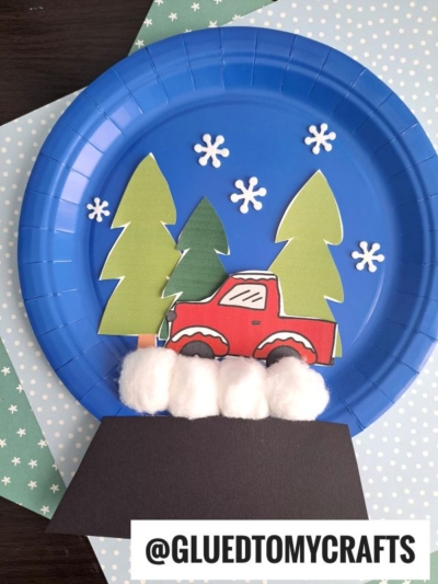 A blue paper plate decorated with green trees, a red truck, white snowflakes, and cotton balls as snow creates a charming Christmas snow globe scene. Finished with a black paper strip and the handle @GLUEDTOMYCRAFTS.