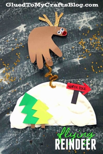 A child’s craft of a paper flying reindeer made from a brown handprint with googly eyes, antlers, and a gold pipe cleaner tail, soars over a paper igloo and North Pole sign with scattered gold stars. Text reads “Flying Reindeer.”.