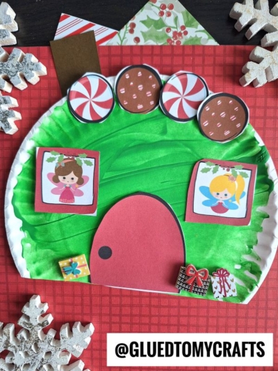 A paper plate Christmas fairy house is crafted in green with a red door, candy decorations, two fairies peeking from window frames, and wrapped gifts—all set on a red checkered background with snowflake ornaments.