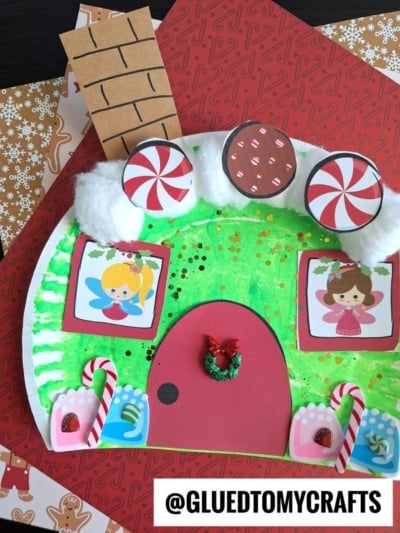 A handmade paper plate Christmas fairy house decorated with cotton balls, candy cutouts, and two fairy illustrations on green and red paper, featuring a chimney and Christmas-themed accents. @GLUEDTOMYCRAFTS is written at the bottom.