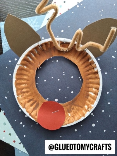 A paper plate reindeer picture frame is painted brown with a cut-out center for your photo, decorated with brown paper ears, a red nose, and twisted pipe cleaner antlers. Snowflake-patterned paper is underneath. Text reads @GLUEDTOMYCRAFTS.