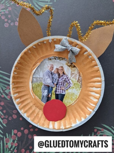 A paper plate reindeer picture frame made with brown paper ears, gold pipe cleaner antlers, a red nose, and silver bow. This adorable reindeer holds a smiling couple's photo in the center. @GLUEDTOMYCRAFTS at the bottom.