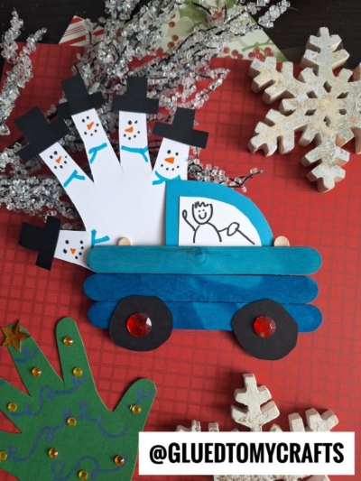 Handmade winter craft featuring a popsicle stick truck decorated as a blue car, with handprint snowmen in the back, snowflakes, and a green handprint tree—all displayed on a red plaid surface. Perfect for popsicle stick Christmas trucks projects!.