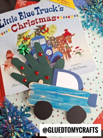 A blue popsicle stick holiday truck craft with a green handprint tree and red gems sits atop the Little Blue Truck's Christmas book, surrounded by snowflake decorations. The image includes the text @GLUEDTOMYCRAFTS.