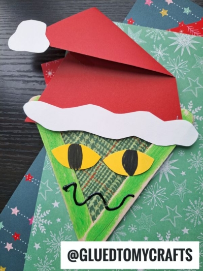 A green, triangular Popsicle Stick Grinch craft with yellow eyes, a wavy black mouth, and a red Santa hat sits on festive holiday paper. The craft resembles the Grinch. @GLUEDTOMYCRAFTS is written at the bottom.