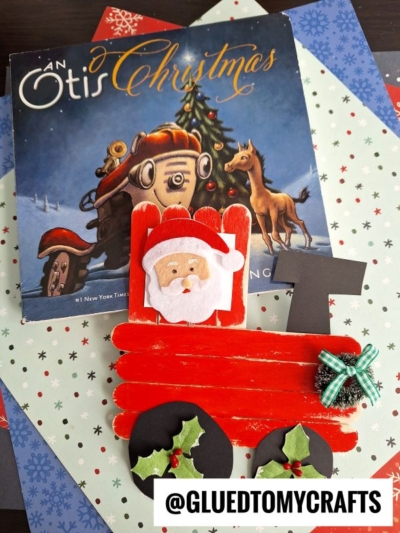 A children’s book titled An Otis Christmas is displayed behind a red popsicle stick craft shaped like a festive tractor with a Santa face, holly decorations, and a green bow—perfect inspiration for popsicle stick Christmas trains. The items rest on snowy-patterned paper.