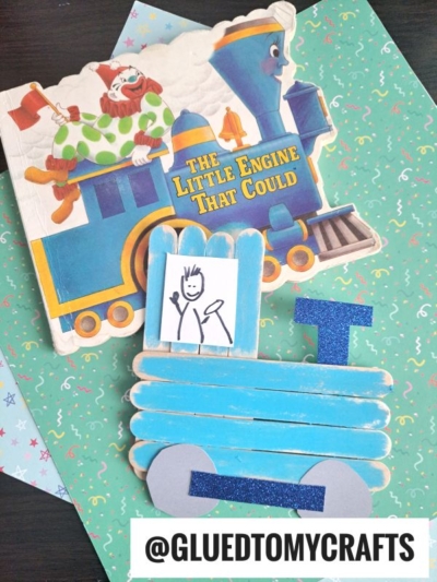 A children’s book titled The Little Engine That Could with a blue train and clown on the cover, next to popsicle stick Christmas trains and a stick-figure drawing of a person placed in the window.