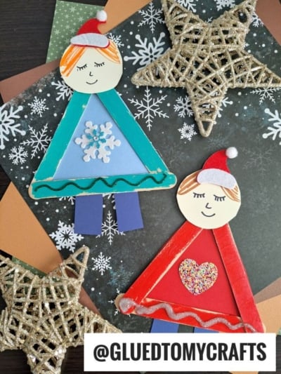 Two handmade Christmas angel crafts with triangle bodies, smiling faces, and red or blue outfits reminiscent of an ugly sweater craft are displayed on festive paper with gold glittery star decorations. @GLUEDTOMYCRAFTS is written at the bottom.