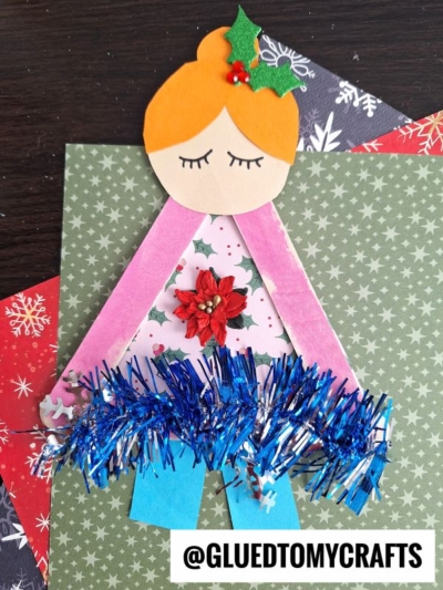 A handmade Christmas ballerina craft using paper, popsicle sticks, tinsel, and festive embellishments—plus a popsicle stick ugly sweater—displayed on snowflake and ornament-patterned paper. @GLUETOMYCRAFTS is printed at the bottom.